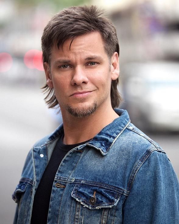 Comedian Theo Von posing outdoors while sporting his signature modern mullet haircut and wearing a classic blue denim jacket over a black shirt.